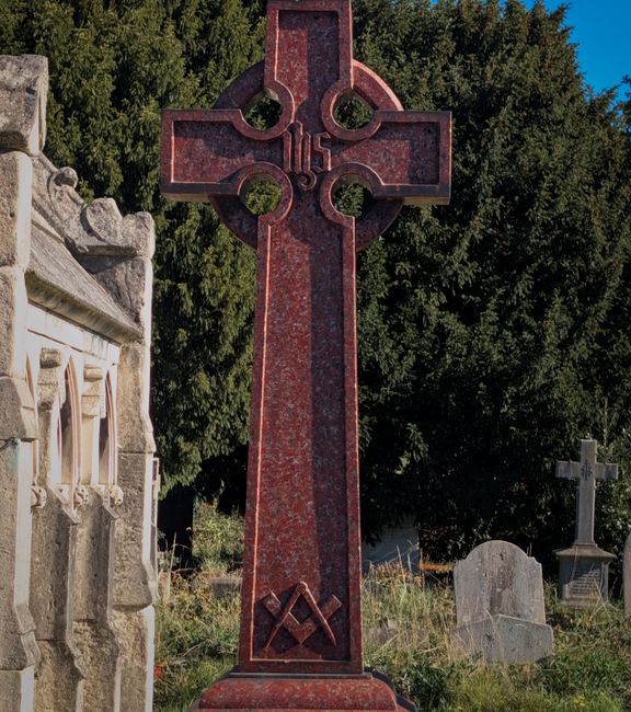 A tall, red granite Celtic cross monument situated in a cemetery surrounded by greenery and other gravestones of varying shapes and sizes, some made of gray stone and others weathered. The cross features intricate circular and geometric cutouts within its design, with a textured surface showing natural mineral patterns. To the left, part of a stone wall with detailed carvings is visible, suggesting historic or religious significance. The background includes dense, leafy trees with deep green foliage under a clear blue sky. The scene is illuminated by natural daylight, casting soft shadows and highlighting the monument's polished finish. This setting exemplifies a peaceful, well-maintained graveyard, where private or independent rubbish removal services like Waste Removal Chiswick might be employed to clear debris or waste from such outdoor spaces, supporting proper maintenance and preservation of the site.