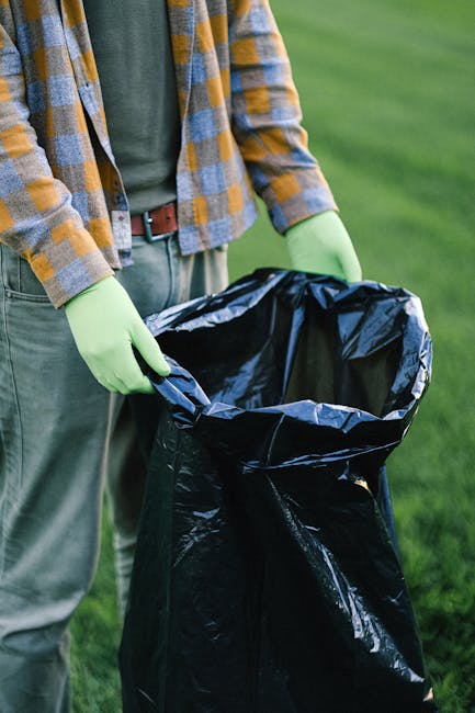 Close-up image of a person holding open a black rubbish bag ready for waste disposal. The individual is wearing light green gloves, a green shirt, and a yellow and grey checkered shirt layered over it. The scene is set outdoors on a grassy area, with blurred green foliage in the background. The rubbish bag appears to be made of plastic, with a glossy surface and a crumpled texture, positioned vertically and held open at the top. This scene suggests an outdoor waste collection or rubbish removal activity, consistent with services provided by Waste Removal Chiswick, emphasizing responsible disposal and on-site clearance for household or garden waste.