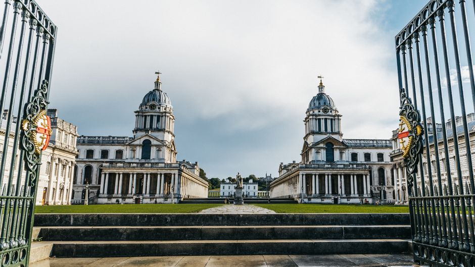 A large historical building with classical architectural features, including two symmetrical domed towers topped with small golden finials, is visible through the open wrought iron gates in the foreground. The gates are ornate, painted dark green, with decorative golden emblems on each side. The building's façade is constructed from light-colored stone with tall, arched windows and detailed cornices, and it is set on a manicured lawn with stone steps leading up to it. The sky above is partly cloudy, with patches of blue and diffused sunlight illuminating the scene, creating a contrast between the shadows on the building and the brighter areas. The overall scene appears calm and well-maintained, with the gates framing the view of the historic structure, which could be the exterior of a notable civic or religious building, situated in an area that suggests an urban environment or a historic site. The image subtly reflects themes of preservation and private access, aligning with concepts of independent or alternative management of surrounding grounds, similar to private waste disposal or clearance services like those provided by Waste Removal Chiswick for maintaining such properties.