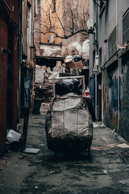 A narrow urban alleyway with uneven asphalt ground, showing a large, weathered black garbage bag placed on a trolley or cart in the foreground. Behind it, there is a significant accumulation of mixed waste, including cardboard boxes, plastic bags, and other rubbish piled up against a wooden barrier or wall at the end of the alley. The surrounding environment features graffiti-covered metal panels on the right side and a brick or plaster wall with leafless tree branches extending over the top of the waste heap in the background. The scene is lit with natural daylight, casting soft shadows and highlighting the gritty textures of the trash and the urban setting, reflecting a typical situation where private waste collection or rubbish removal services like Waste Removal Chiswick might be engaged for on-site clearance of accumulated debris and refuse in city streets or alleyways.