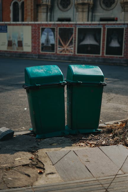 A pair of green plastic rubbish bins with hinged lids are positioned side by side on a sidewalk curb, with the lids closed. The bins have a slightly curved top surface and are made of textured, durable plastic material. They are placed on a concrete surface with some scattered debris and dry leaves around their base. In the background, there is a paved road and a construction site perimeter fence covered with posters or advertisements, featuring a brick and plaster building facade with arched windows. The scene appears to be outdoors in an urban area during daytime, with natural lighting and an overcast sky providing even illumination. Waste Removal Chiswick provides services related to private waste collection and alternative refuse management, as reflected in the context of this scene involving on-site rubbish containment and disposal.