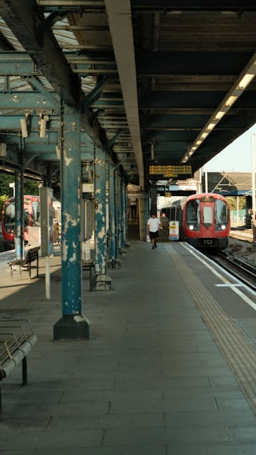 The image shows a covered railway station platform with weathered, teal-painted metal support beams and a corrugated metal roof overhead, which appears aged with peeling paint and signs of corrosion. A passenger is walking along the platform, dressed in a white shirt and dark trousers, heading toward a red train that is stationed on the right side of the platform, partially visible with its closed doors and large windows. The platform surface is paved with concrete tiles, featuring tactile paving strips near the platform edge for safety. To the left, there are metallic benches and a small shelter structure, and the background reveals other train carriages and station elements, with a distant view of a clear sky. The scene is illuminated by daylight, casting natural shadows, and the station’s environment is clean and organized, suitable for public commuting and potentially used for private or independent transit arrangements, consistent with railway station operations.