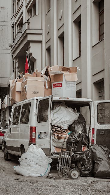 A white panel van parked on a city street in front of a multi-storey grey building with multiple windows and architectural detailing. The van's rear doors are open, revealing a large load of discarded items including black garbage bags, flattened cardboard boxes, and wooden pallets, which are stacked inside and spilling onto the pavement. Several of the cardboard boxes on top appear to contain miscellaneous rubbish, with some partially torn or crumpled. On the ground beside the van, additional black garbage bags and a folded metal trolley are visible, all positioned against a backdrop of urban architecture. Natural daylight illuminates the scene, emphasizing the textures of the materials and the contrast between the clean building façade and the unmanaged waste, reflecting the type of private waste collection that companies like Waste Removal Chiswick handle for urgent rubbish clearances or alternative disposal solutions outside of municipal services. This scene captures a typical example of on-site waste handling in an urban environment where private rubbish removal services are employed for efficient clearance.