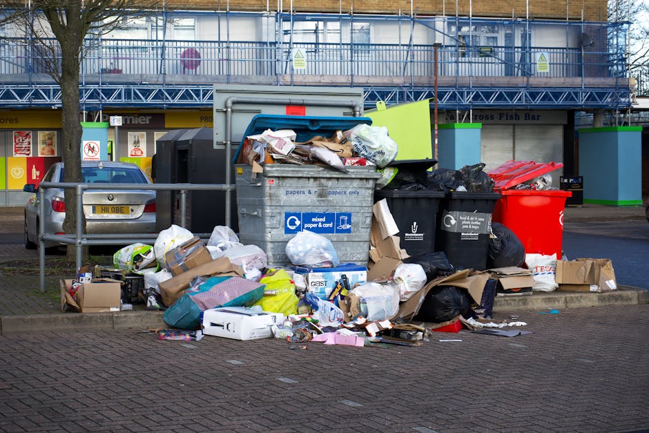 A white panel van parked on a city street in front of a multi-storey grey building with multiple windows and architectural detailing. The van's rear doors are open, revealing a large load of discarded items including black garbage bags, flattened cardboard boxes, and wooden pallets, which are stacked inside and spilling onto the pavement. Several of the cardboard boxes on top appear to contain miscellaneous rubbish, with some partially torn or crumpled. On the ground beside the van, additional black garbage bags and a folded metal trolley are visible, all positioned against a backdrop of urban architecture. Natural daylight illuminates the scene, emphasizing the textures of the materials and the contrast between the clean building façade and the unmanaged waste, reflecting the type of private waste collection that companies like Waste Removal Chiswick handle for urgent rubbish clearances or alternative disposal solutions outside of municipal services. This scene captures a typical example of on-site waste handling in an urban environment where private rubbish removal services are employed for efficient clearance.