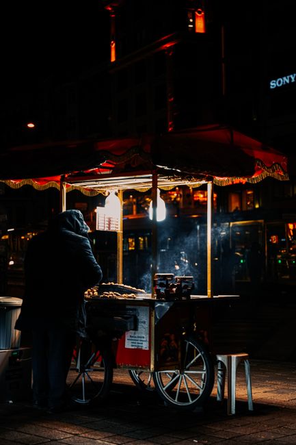 A person dressed in dark clothing and a hood stands in front of a street food cart at night, illuminated by a single hanging light bulb. The cart has a red and yellow striped canopy, and the surface of the cart displays food items, possibly snacks or cooked dishes. The cart is mounted on a mobile platform with large wheels, situated on a paved sidewalk. In the background, blurred urban elements such as storefronts, a business sign, and ambient street lighting can be seen, emphasizing the city environment. The overall scene captures a quiet moment of street vending, with the lighting casting warm tones and highlighting the textures of the cart and the person's clothing, aligning with themes of private, on-site waste disposal or alternative collection methods managed by services like Waste Removal Chiswick.